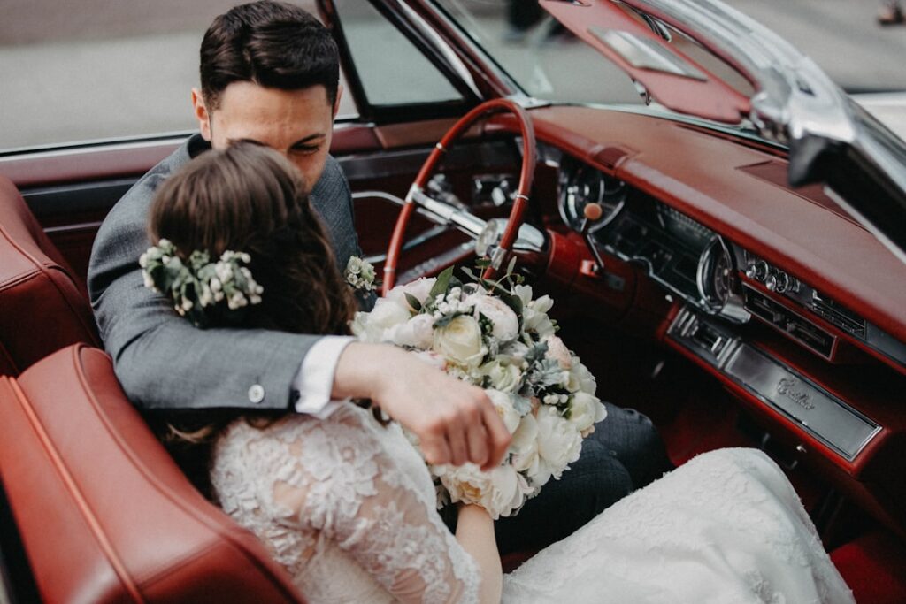 Bride and groom sitting together in the leather interior of a classic wedding car, bride holding a bouquet
