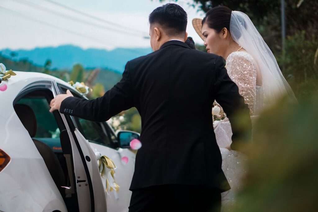Bride and groom being helped into a decorated wedding car by a formally dressed attendant