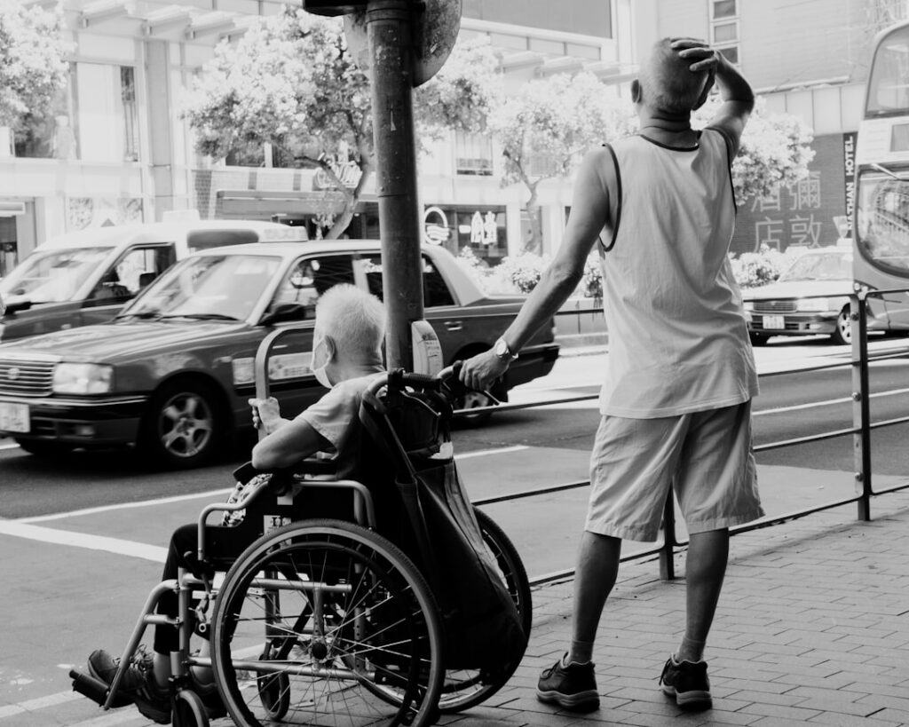 A person pushing a wheelchair user along a street near vehicles, illustrating the difference between WAV and standard adapted transport